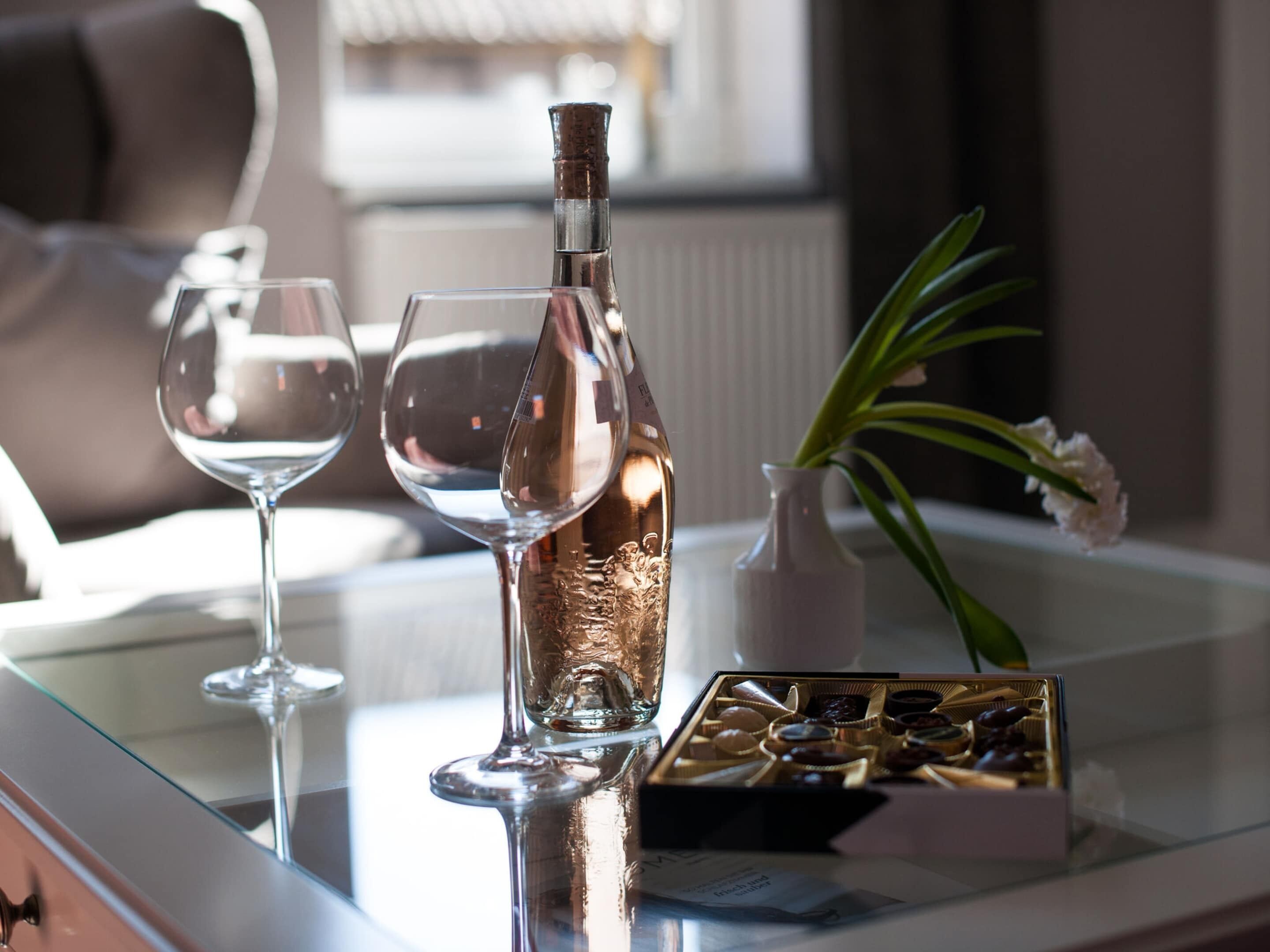 A glass table with a bottle, two wine glasses, a vase with a flower, and a box of chocolates.