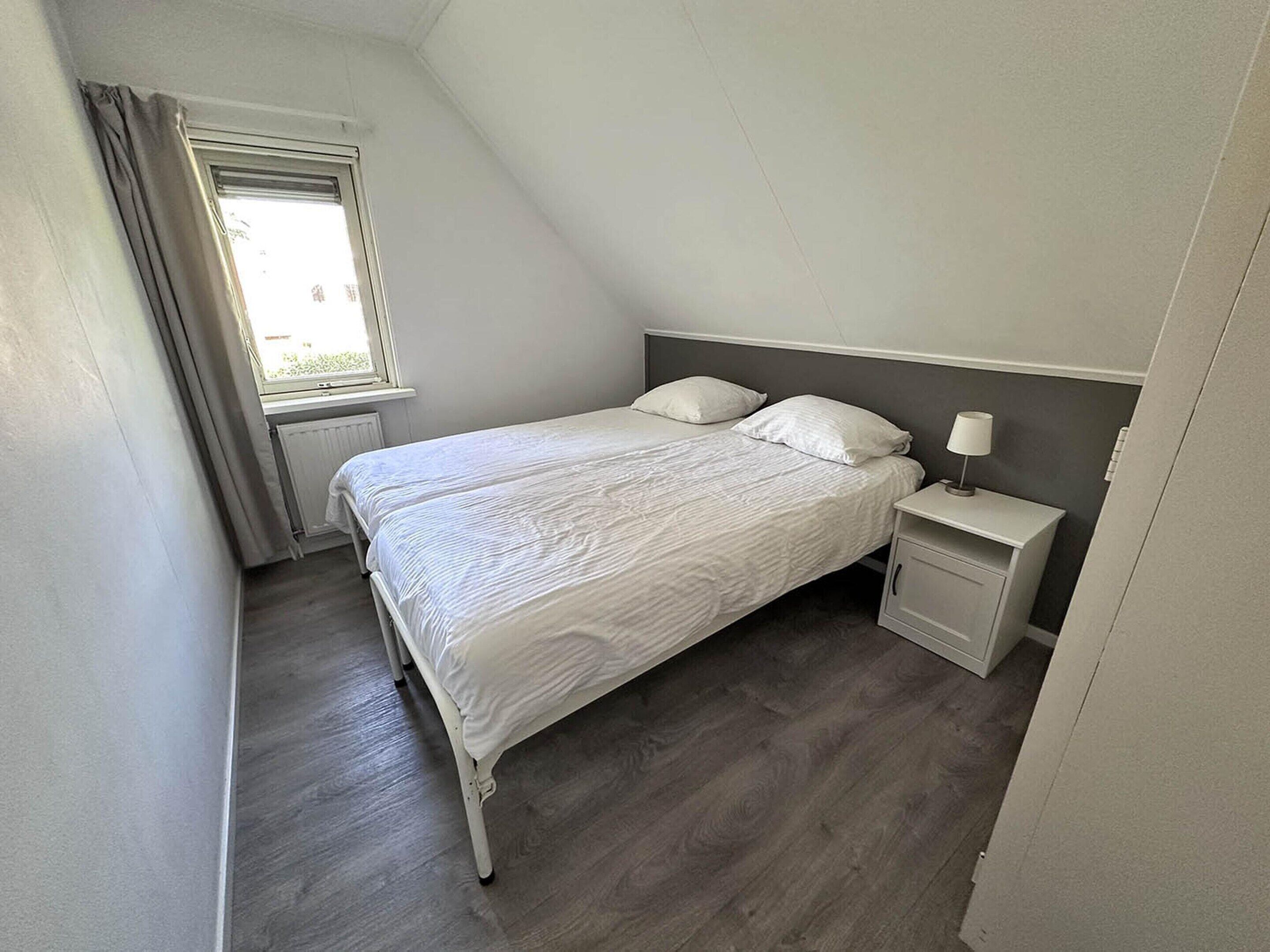 Una cama individual en una habitación con techo inclinado, una mesita de noche con lámpara y una ventana con cortinas.