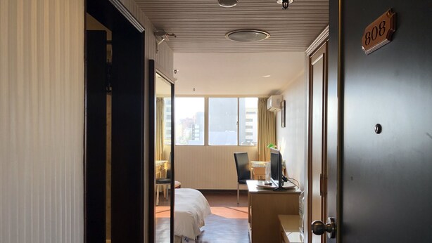 A hotel room with a bed, a desk with a computer, a chair, and a view of the city through large windows. - Hermes Hotel (Hsinchu)