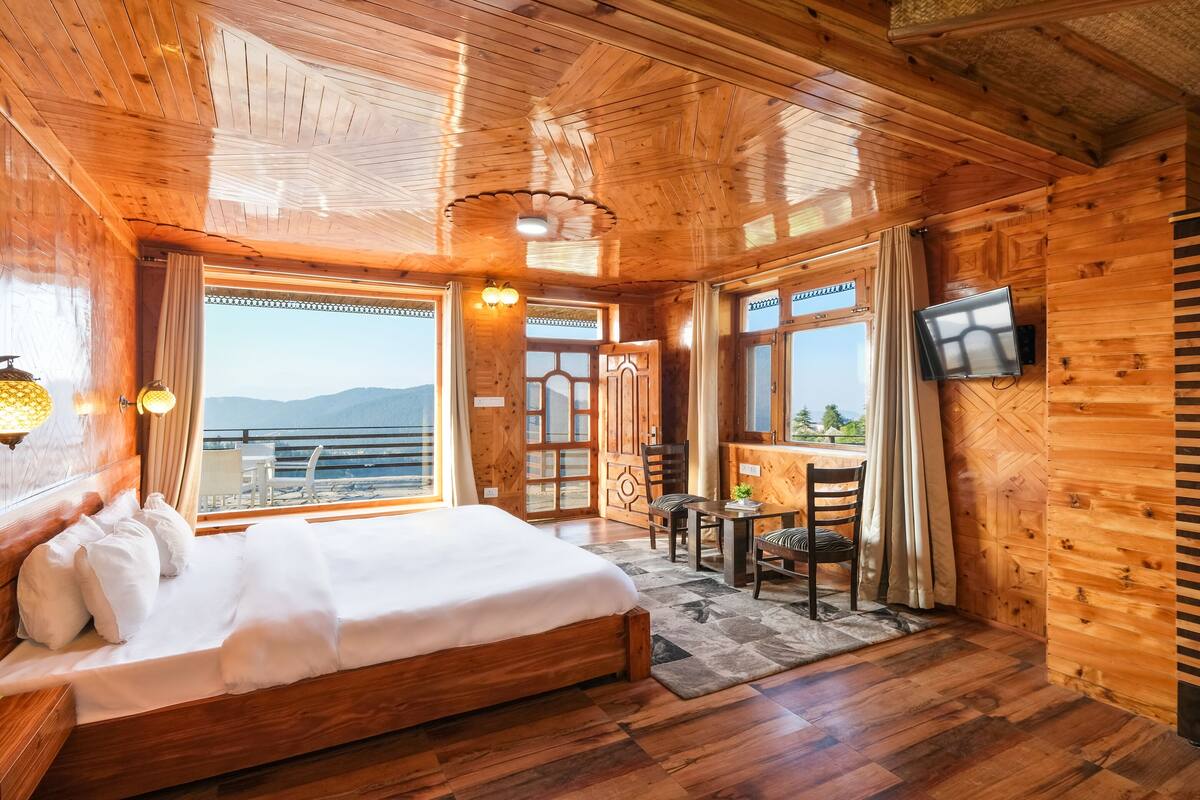 Habitación de cabaña de madera con cama, mesa de comedor, sillas, televisor y ventanas amplias con vistas a las montañas.