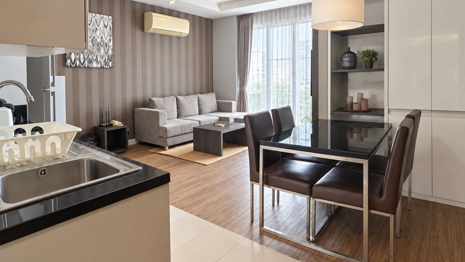 A modern kitchen with a built-in sink, a dining area with a glass table and chairs, a grey sofa, and a large window with curtains.