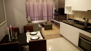 Eine moderne Küche mit weißen Schränken, einer schwarzen Arbeitsplatte und Edelstahlgeräten. Es gibt einen Essbereich mit einem Tisch für zwei Personen, einer Couch und einem Couchtisch. Der Raum verfügt über ein großes Fenster mit Vorhängen.