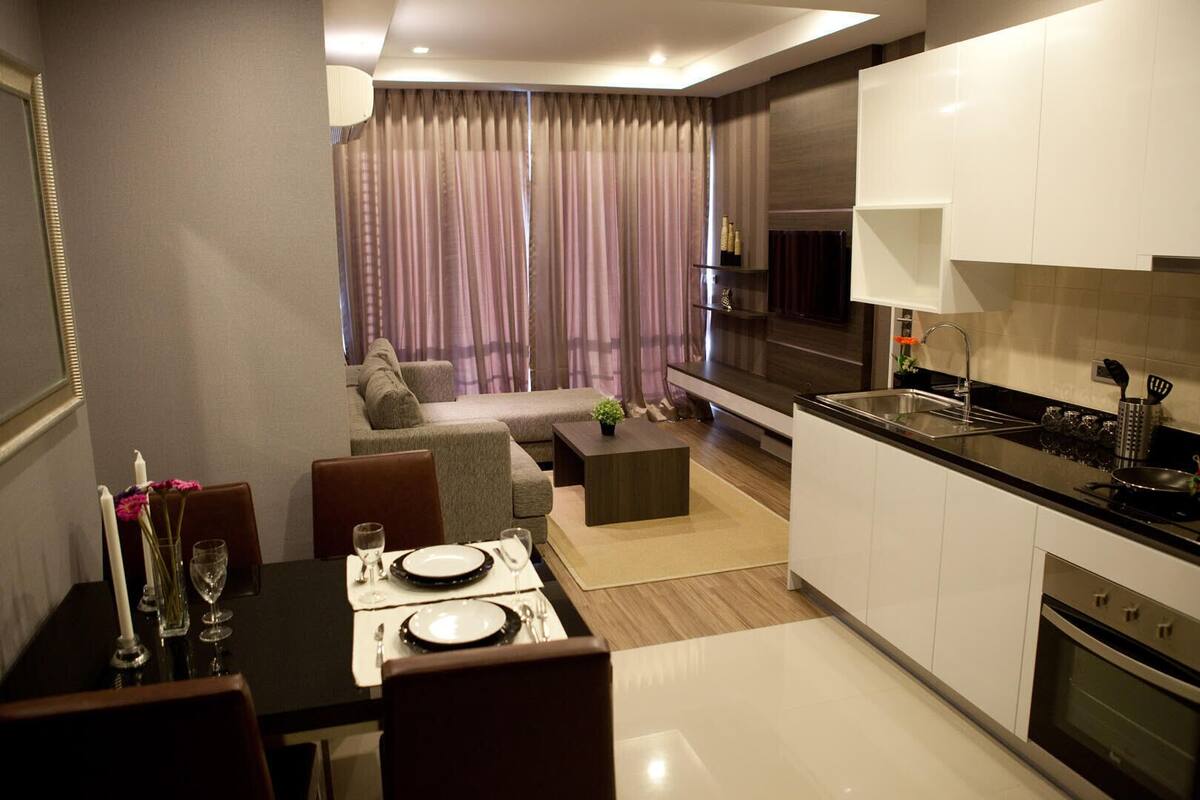 Una cocina moderna con armarios blancos, encimera negra y electrodomésticos de acero inoxidable. Hay una zona de comedor con una mesa preparada para dos personas, un sofá y una mesa de centro. La habitación tiene un ventanal con cortinas.