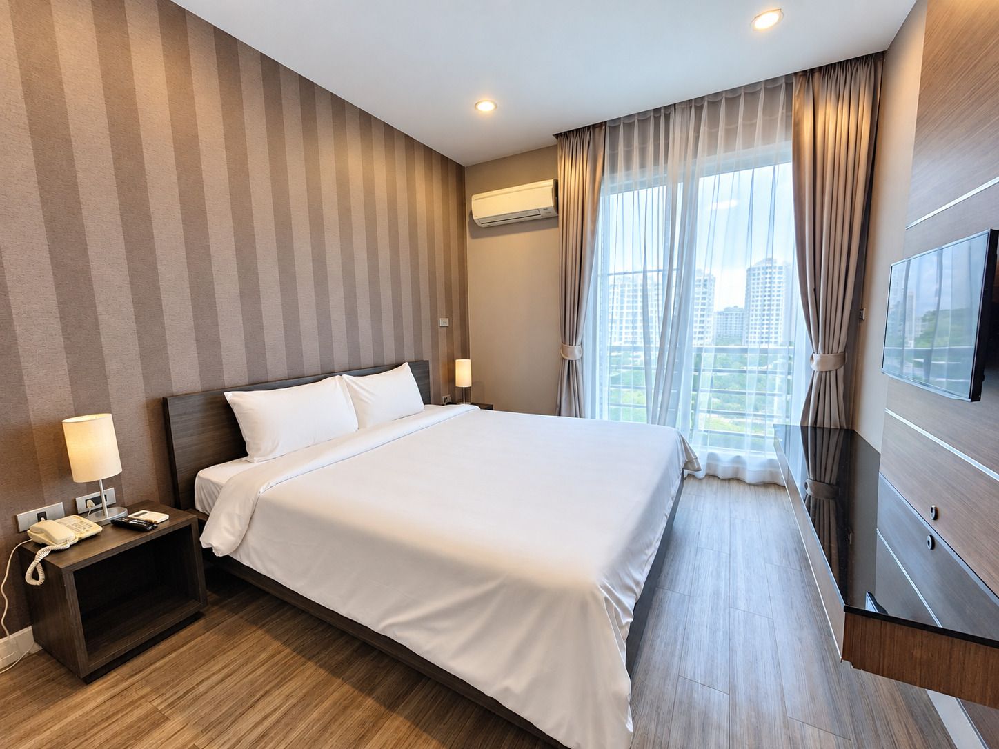 A hotel room with a large bed, a bedside table with a lamp, an air conditioning unit, a TV, and a window with curtains.