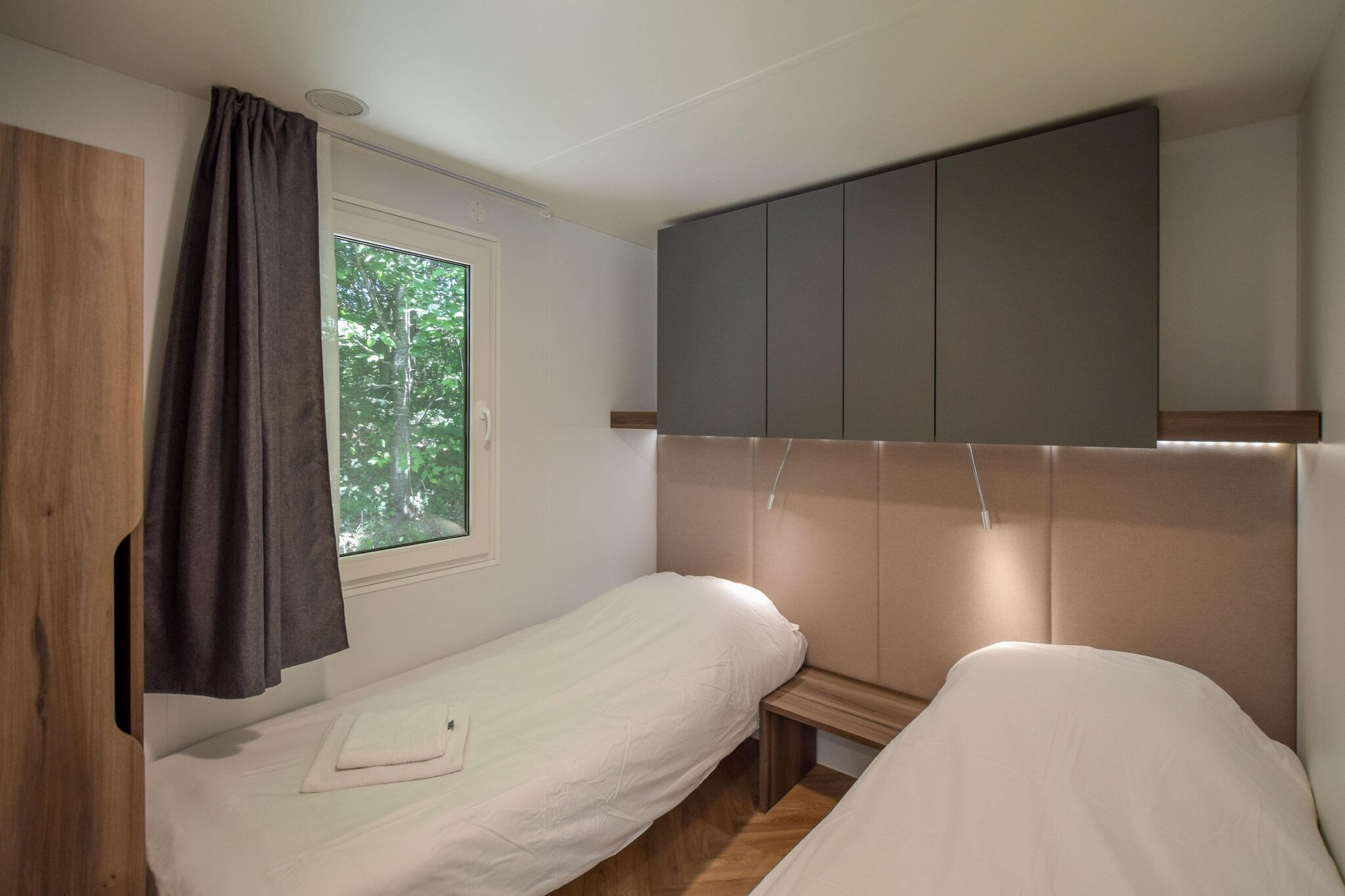 Un dormitorio con dos camas individuales, una ventana con vistas a árboles, un armario de madera y un cabecero gris.