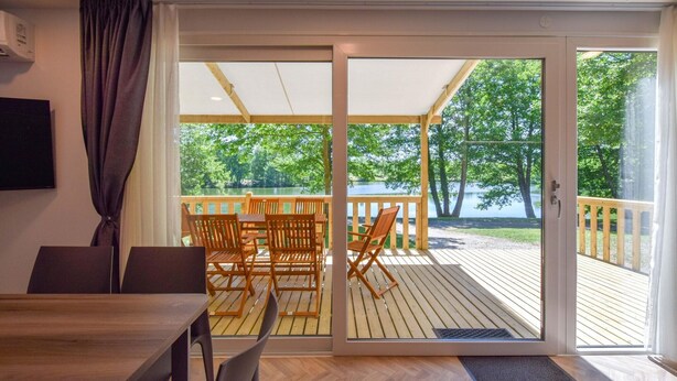 Una habitación con una mesa y sillas de madera, una puerta corrediza de vidrio que da a una terraza exterior con vistas a un lago y árboles.