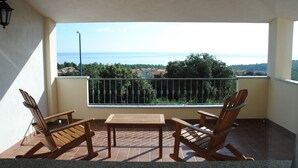 A balcony with two wooden rocking chairs and a table, overlooking a coastal view with houses and greenery.