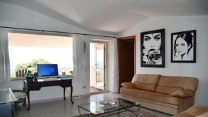 A living room with a glass coffee table, a brown leather sofa, a desk with a computer, and two framed artworks on the wall.