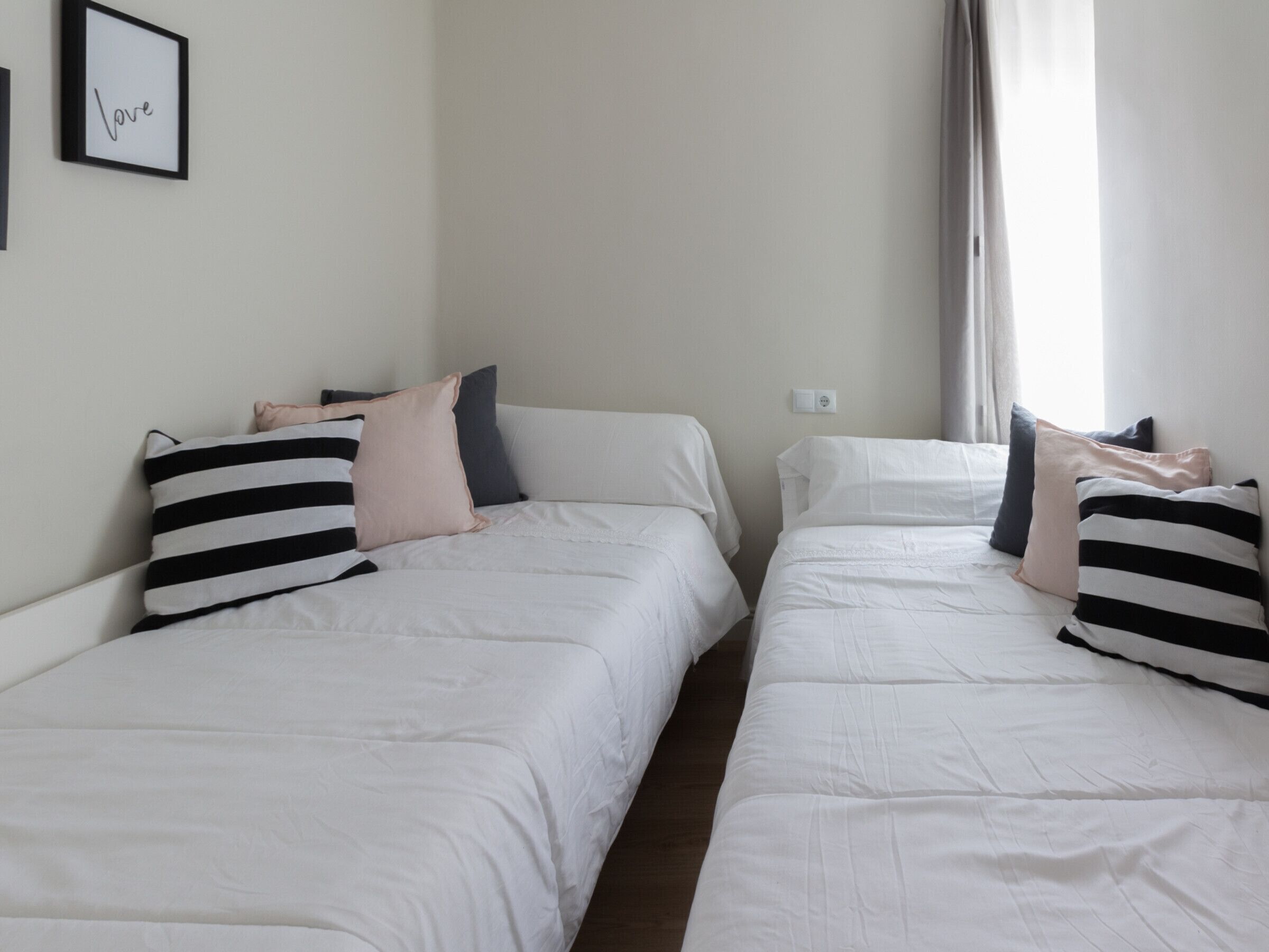 A room with two single beds, each with white bedding and striped pillows. There is a framed picture on the wall and a window with curtains.