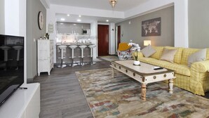 A modern living room with a yellow sofa, a coffee table, a TV, and a kitchen area in the background.