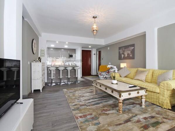A modern living room with a yellow sofa, a coffee table, a TV, and a kitchen area in the background. - Bright apartment with terrace in central Malaga (Málaga)