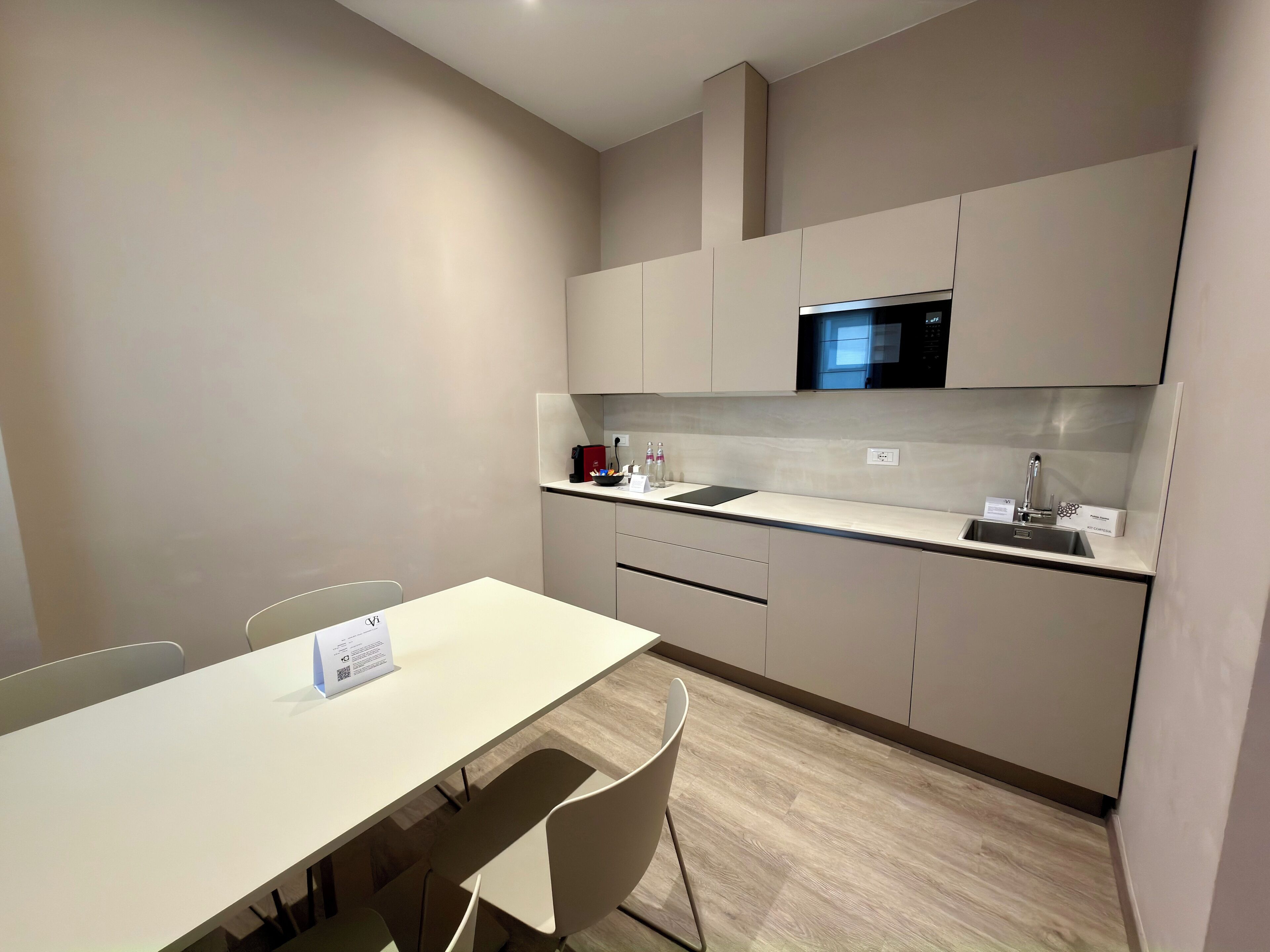 Una cocina moderna con encimera blanca, pileta de acero inoxidable y microondas. Hay una mesa de comedor con sillas y una pequeña señal encima.