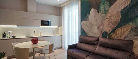 A modern living room with a brown sofa, a round dining table with chairs, a kitchen area with a microwave, and a large leaf-patterned wall mural.