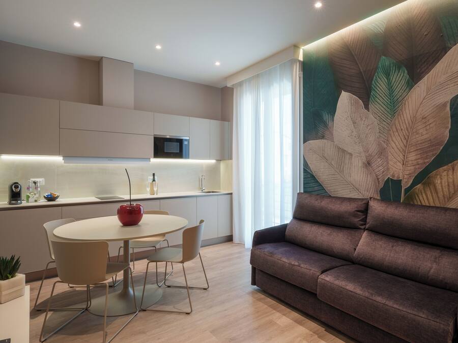 Ein modernes Wohnzimmer mit einer braunen Couch, einem runden Esstisch mit Stühlen, einer Küchenzeile mit Mikrowelle und einer großen Wandmalerei mit Blattmuster.
