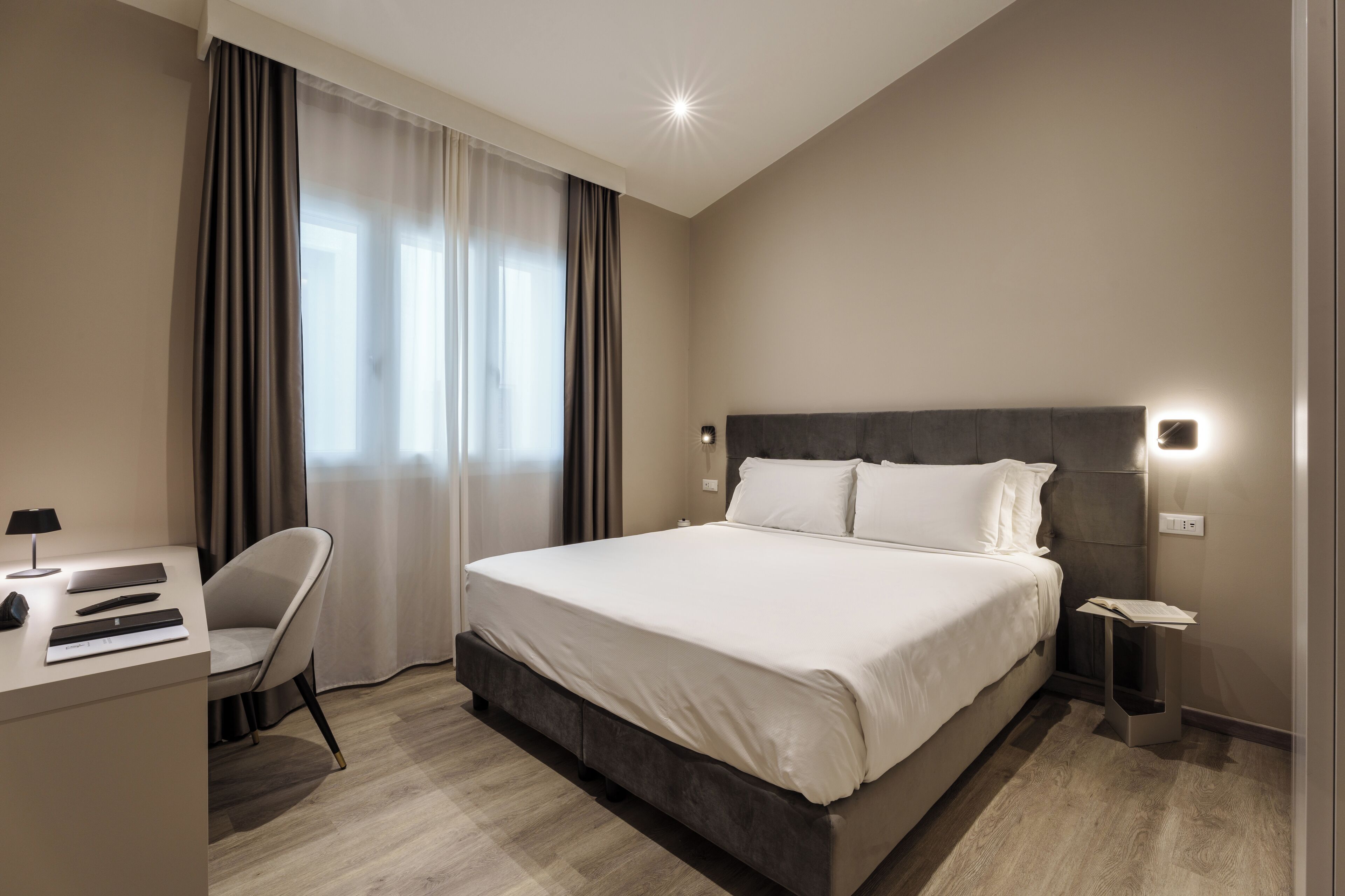 Quarto de hotel moderno com uma cama grande, uma escrivaninha com cadeira e uma mesinha de cabeceira.