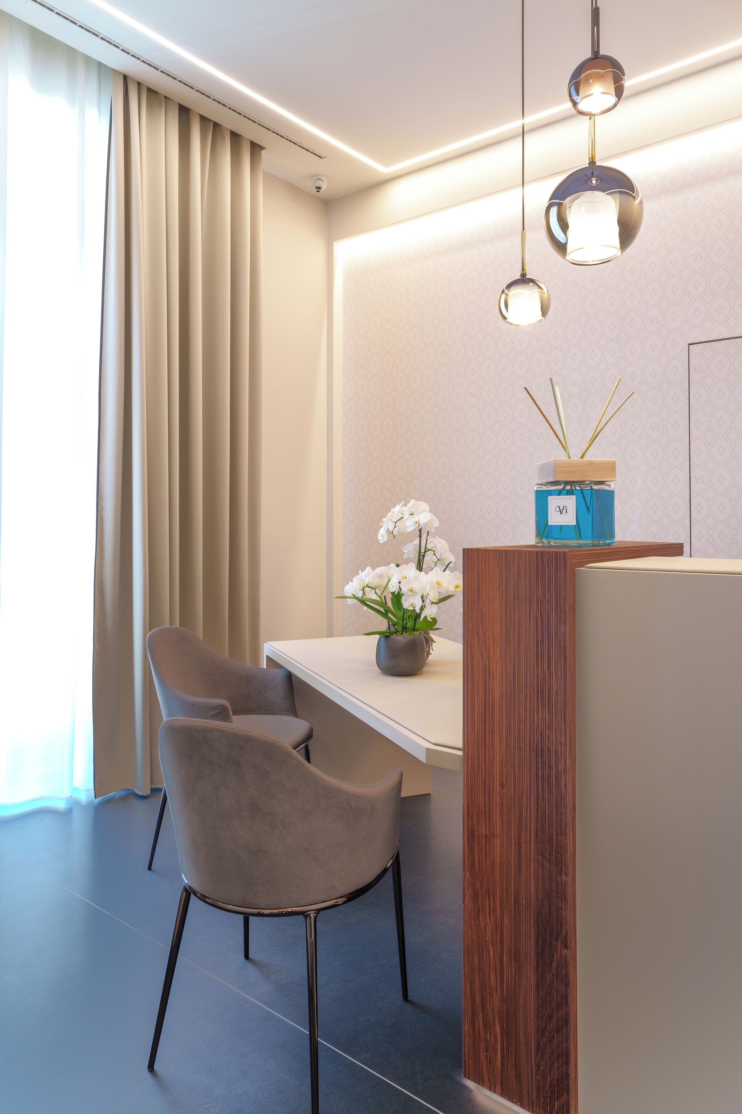 Una habitación de hotel moderna con comedor que presenta una partición de madera, dos lámparas colgantes, un florero con flores blancas y un aparador de madera.