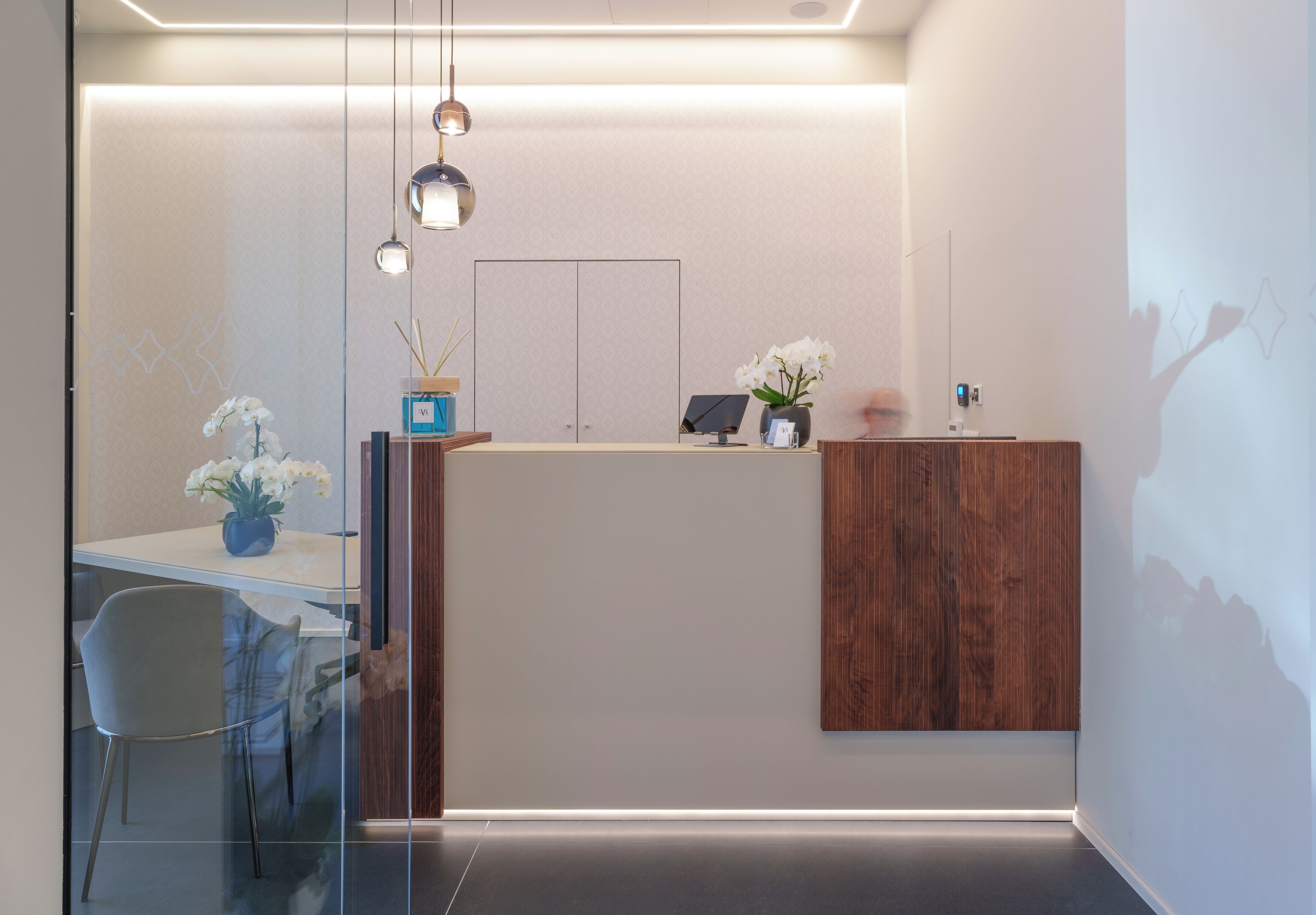 A modern reception area with a glass partition, a wooden reception desk, and a minimalist decor including a vase with white flowers and a small potted plant.