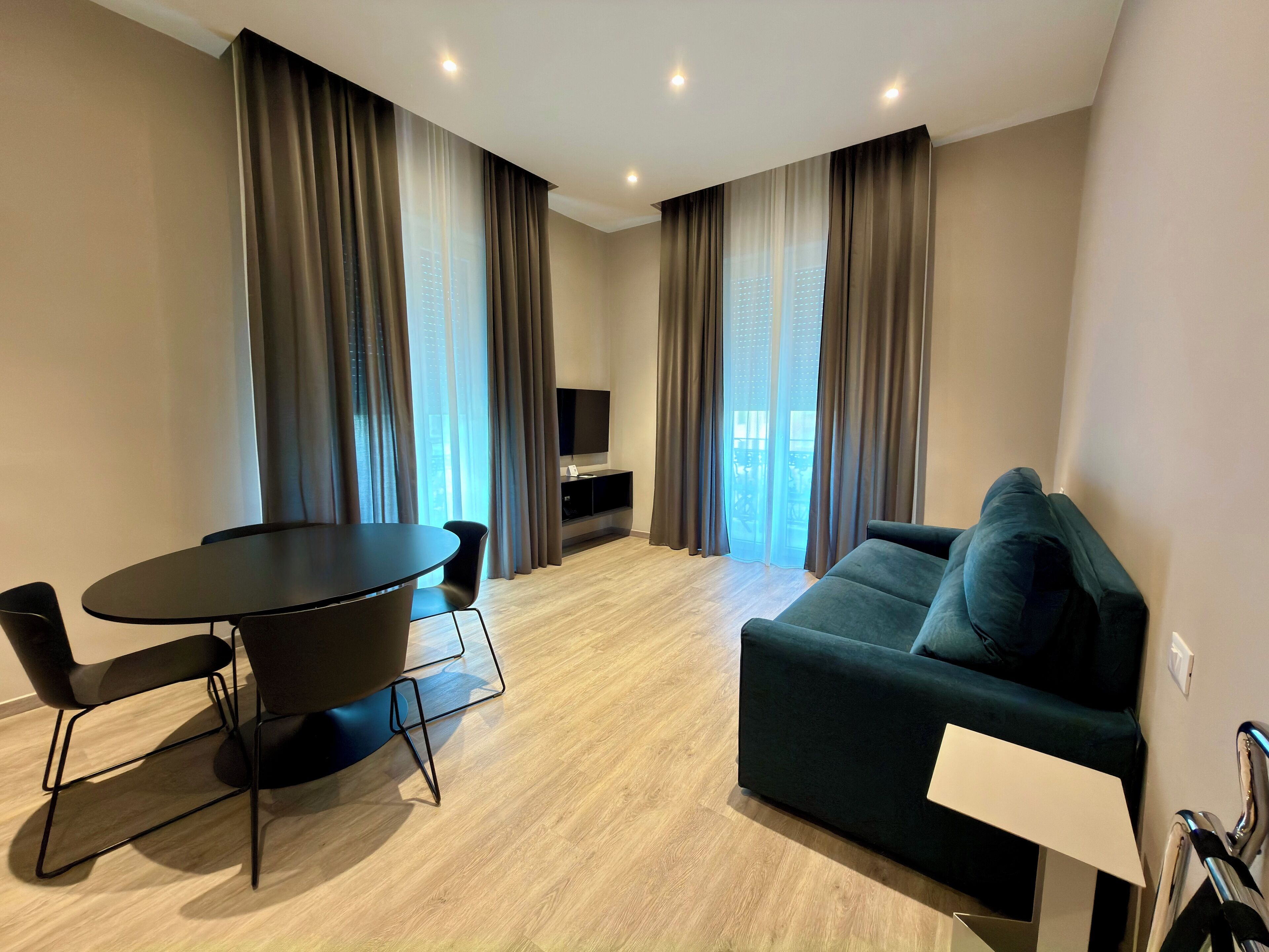 A modern living room with a dark green sofa, a round dining table with chairs, and a flat-screen TV mounted on the wall.