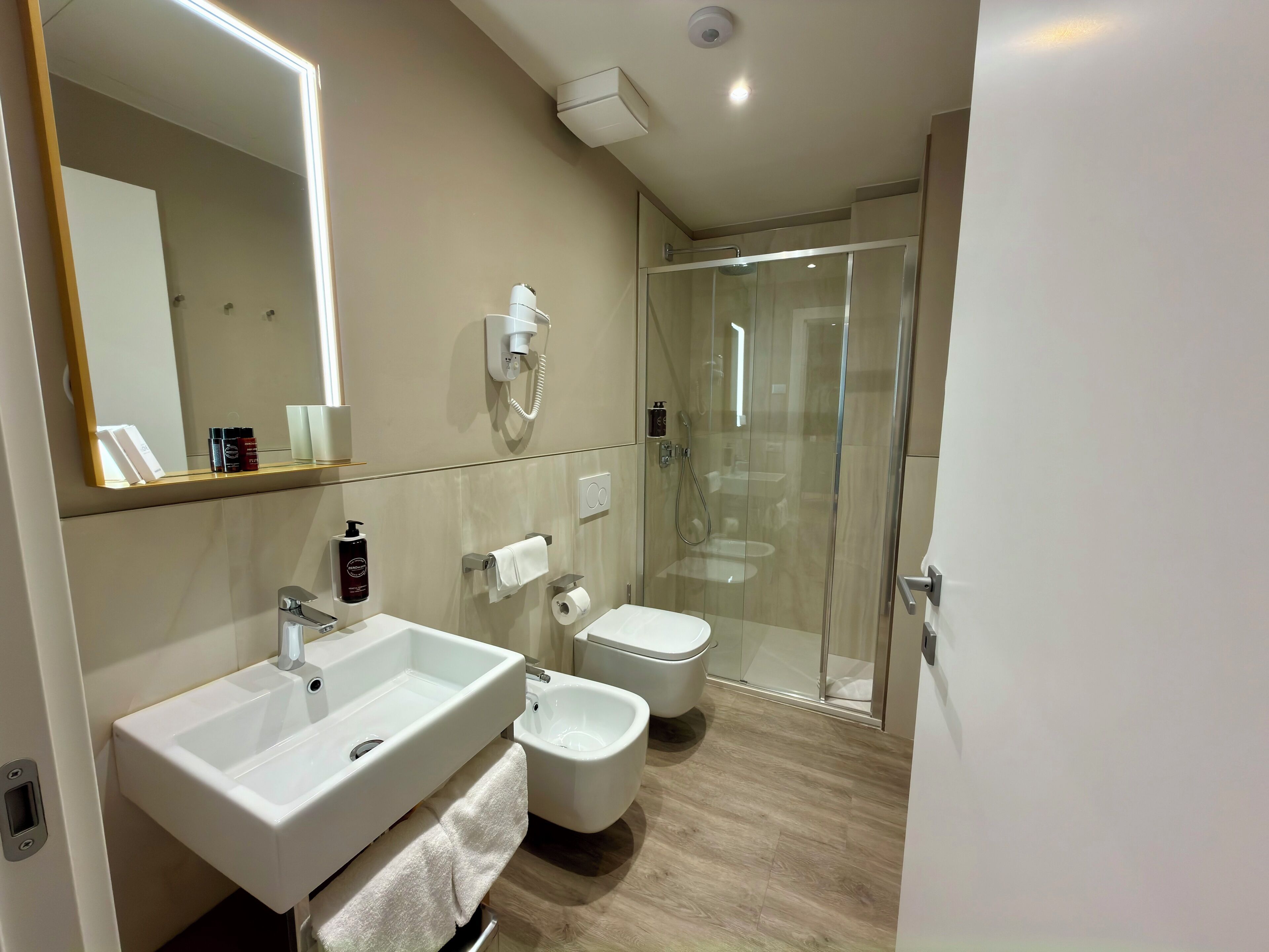 A modern bathroom with two sinks, a bidet, a toilet, a glass shower enclosure, and a mirror with integrated lighting.