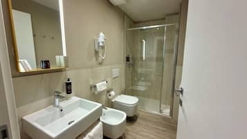 A modern bathroom with two sinks, a bidet, a toilet, a glass shower enclosure, and a mirror with integrated lighting.