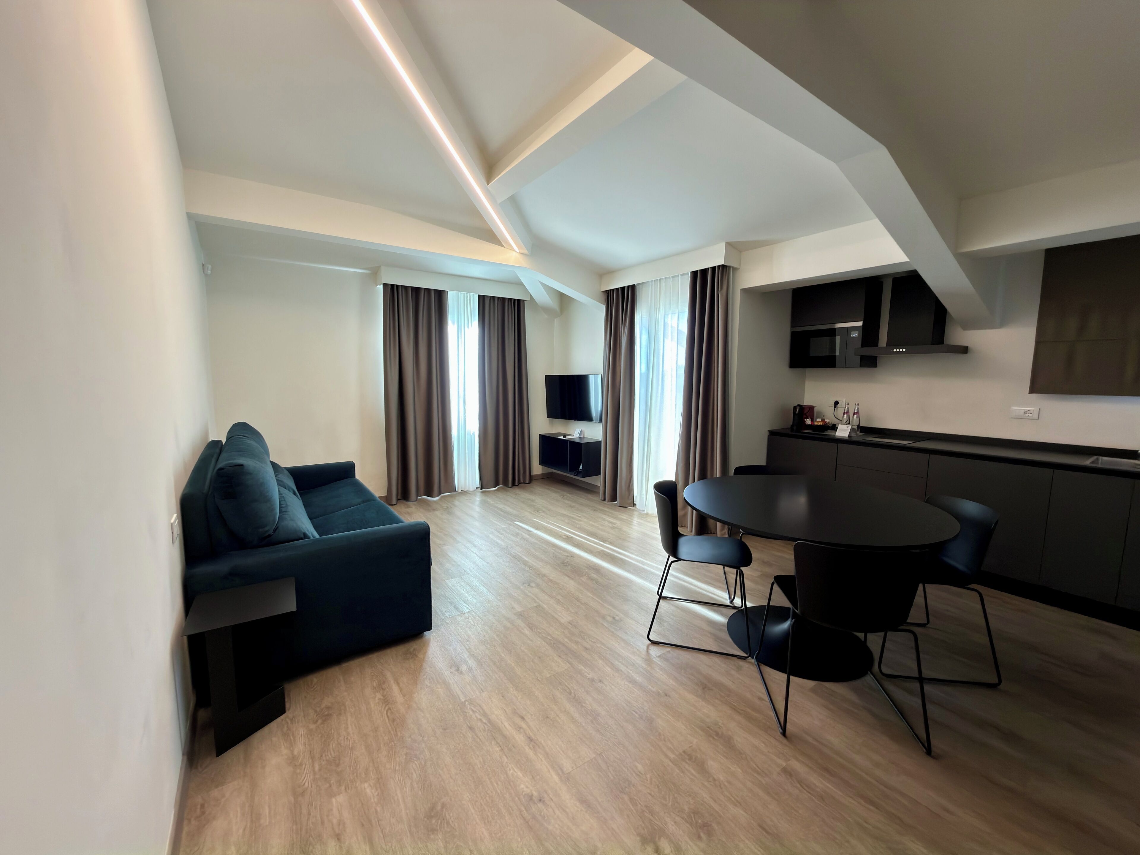 A modern living room with a dark blue sofa, a dining table with chairs, a kitchen area with a microwave, and a TV mounted on the wall.