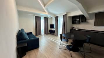A modern living room with a dark blue sofa, a dining table with chairs, a kitchen area with a microwave, and a TV mounted on the wall.