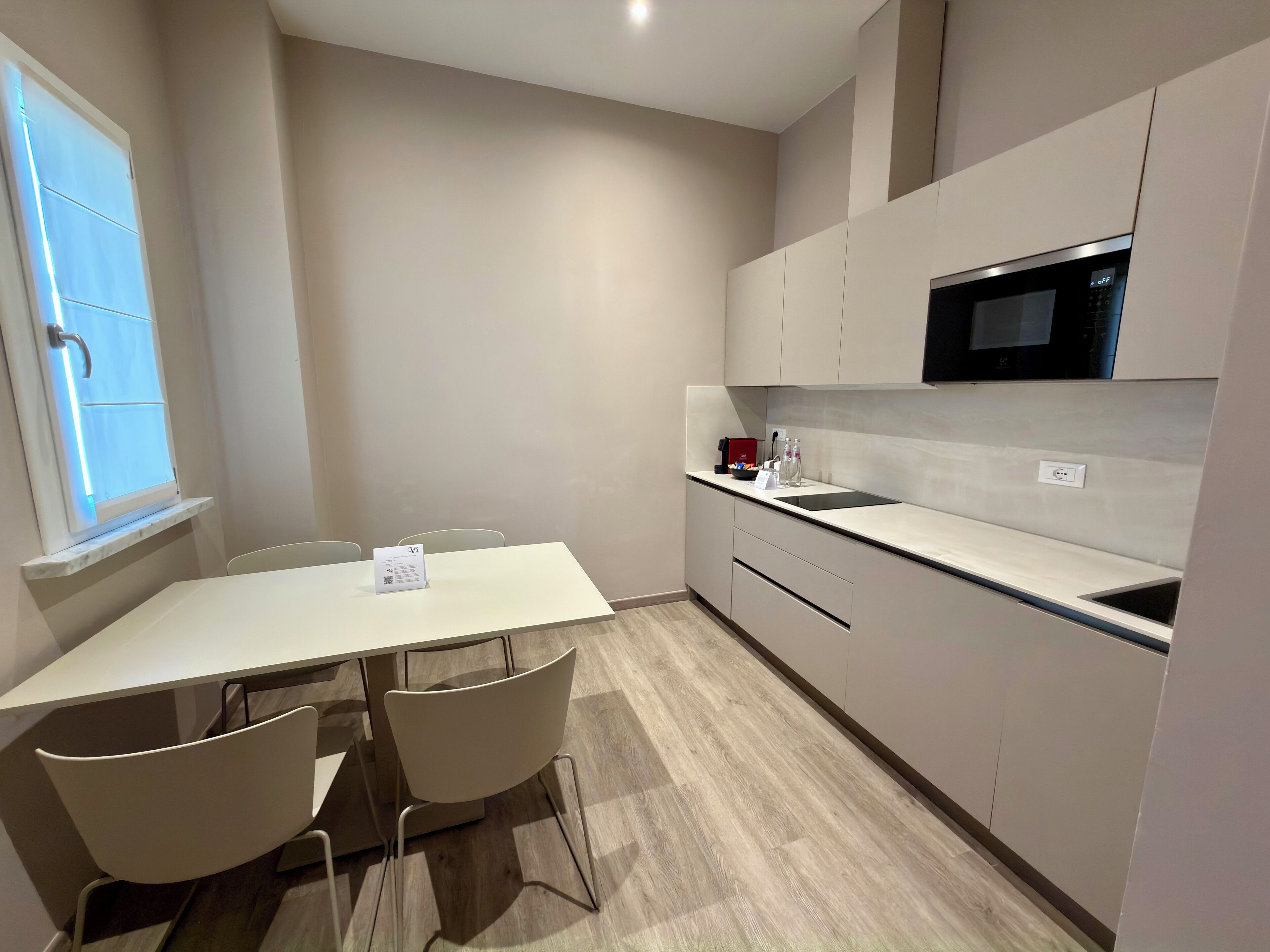 A modern kitchen with a white countertop, a microwave, and a sink. There is a dining table with chairs and a window with blinds.