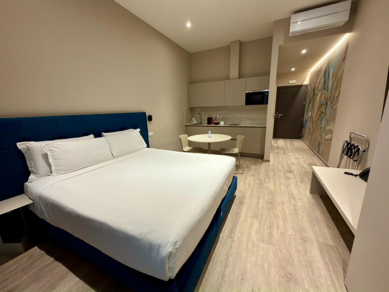 A modern hotel room with a large bed, a small kitchenette, a dining area, and a bathroom.