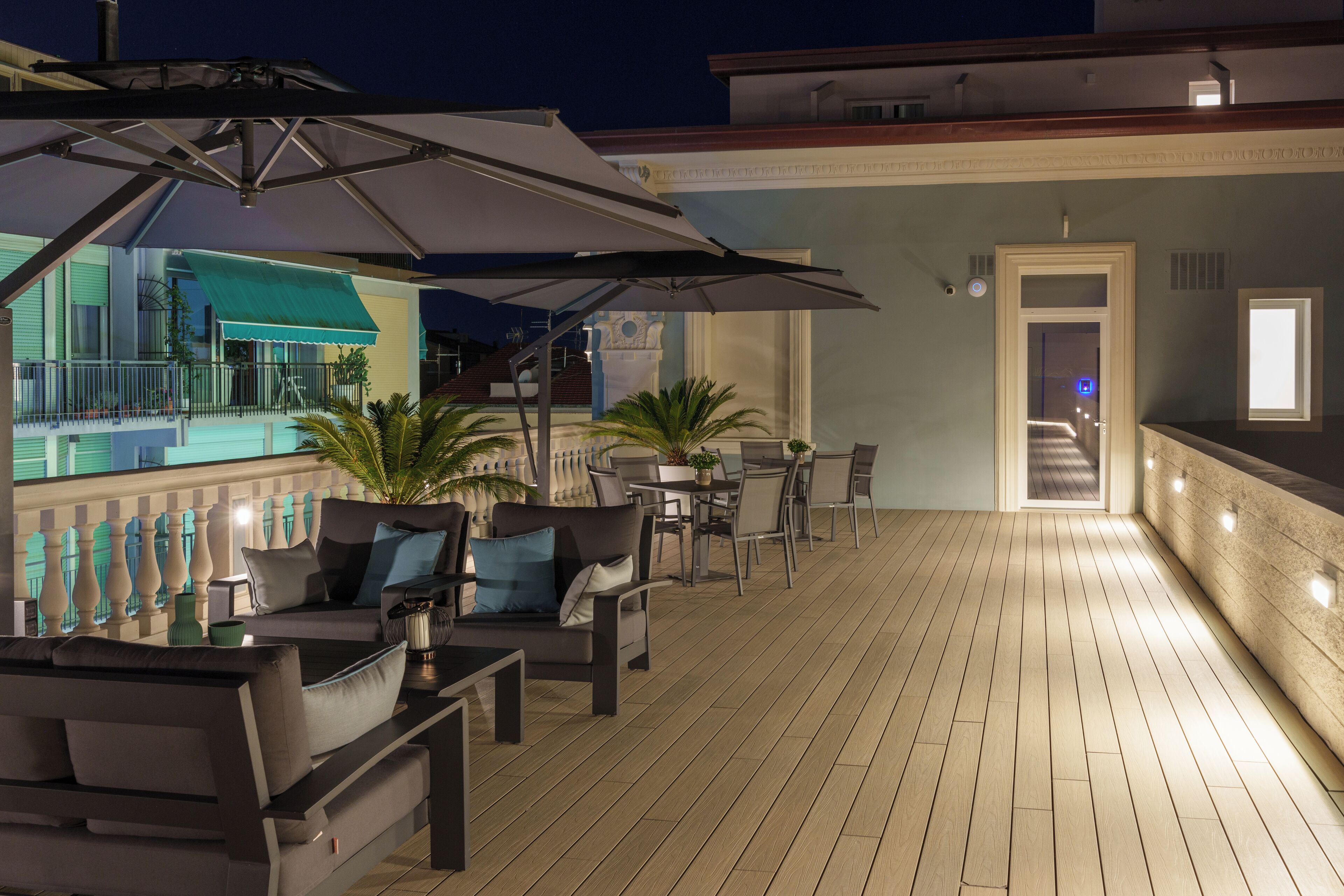 Um deck ao ar livre com piso de madeira, mobiliário externo, guarda-sóis e uma piscina ao fundo.