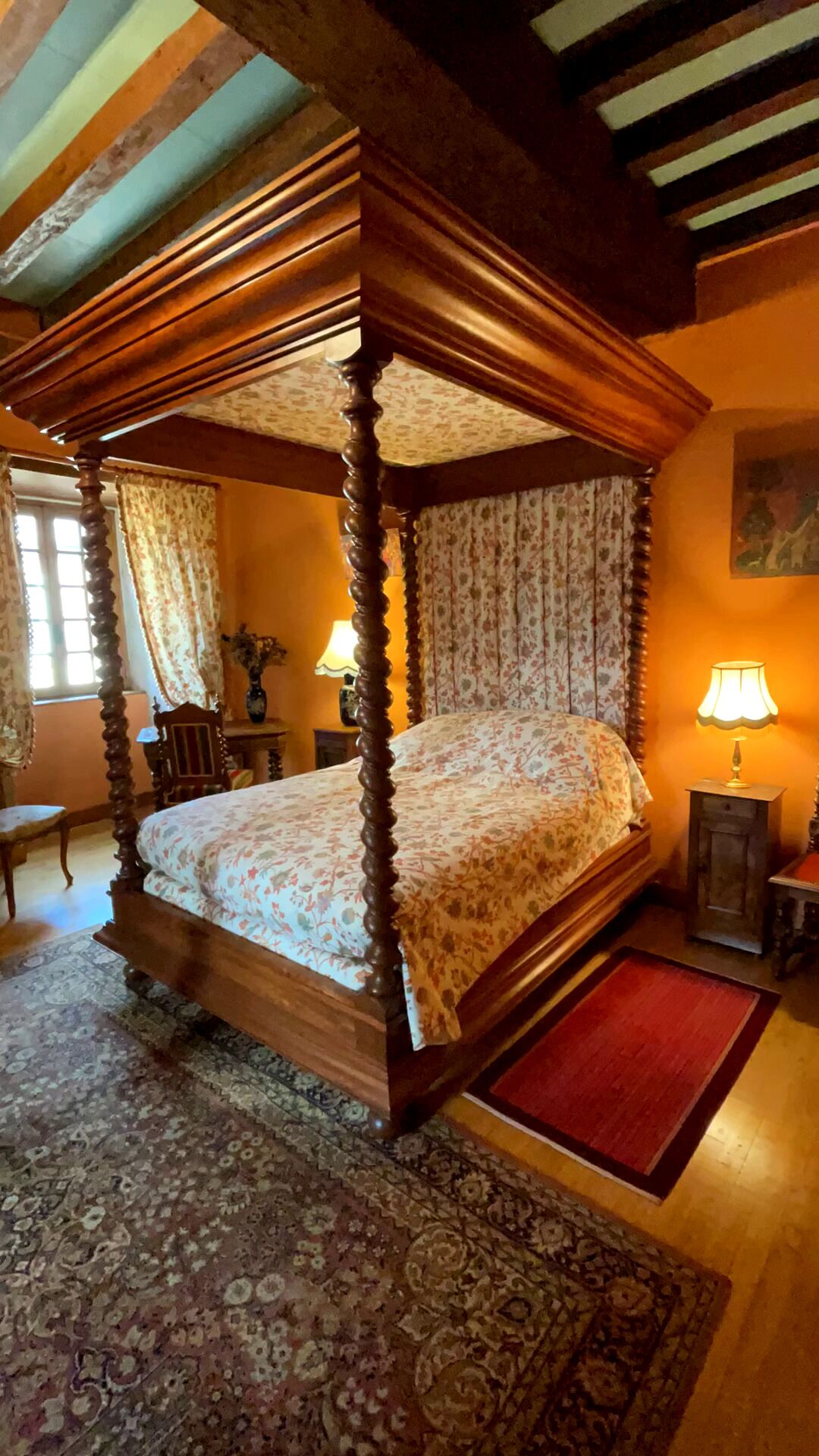 Een hemelbed met bloemenbeddengoed, een rood vloerkleed en een klein tafeltje met een lamp.