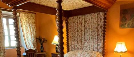 Een hemelbed met bloemenbeddengoed, een rood vloerkleed en een klein tafeltje met een lamp.