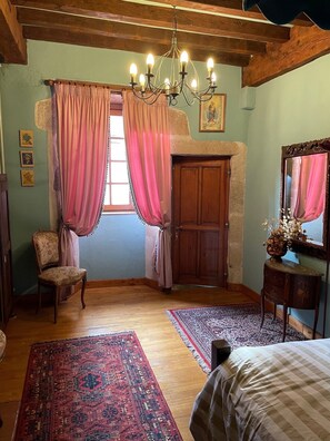 Una habitación con techo de madera, un candelabro, un ventanal con cortinas rosadas, una puerta de madera, una silla, una cama, una mesita con un florero y un espejo.