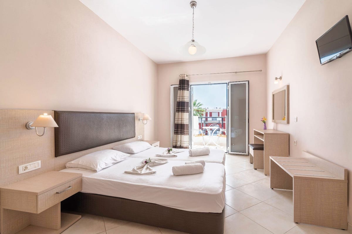 A modern hotel room with a large bed, a bedside table, a TV mounted on the wall, and a balcony with a view of the city.