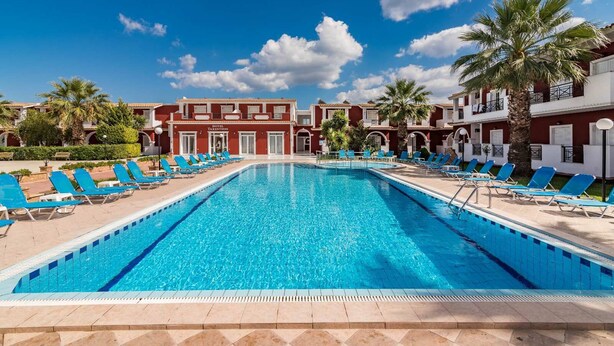 A hotel with a large swimming pool, blue lounge chairs, and palm trees. - Yakinthos Hotel (Zakynthos)