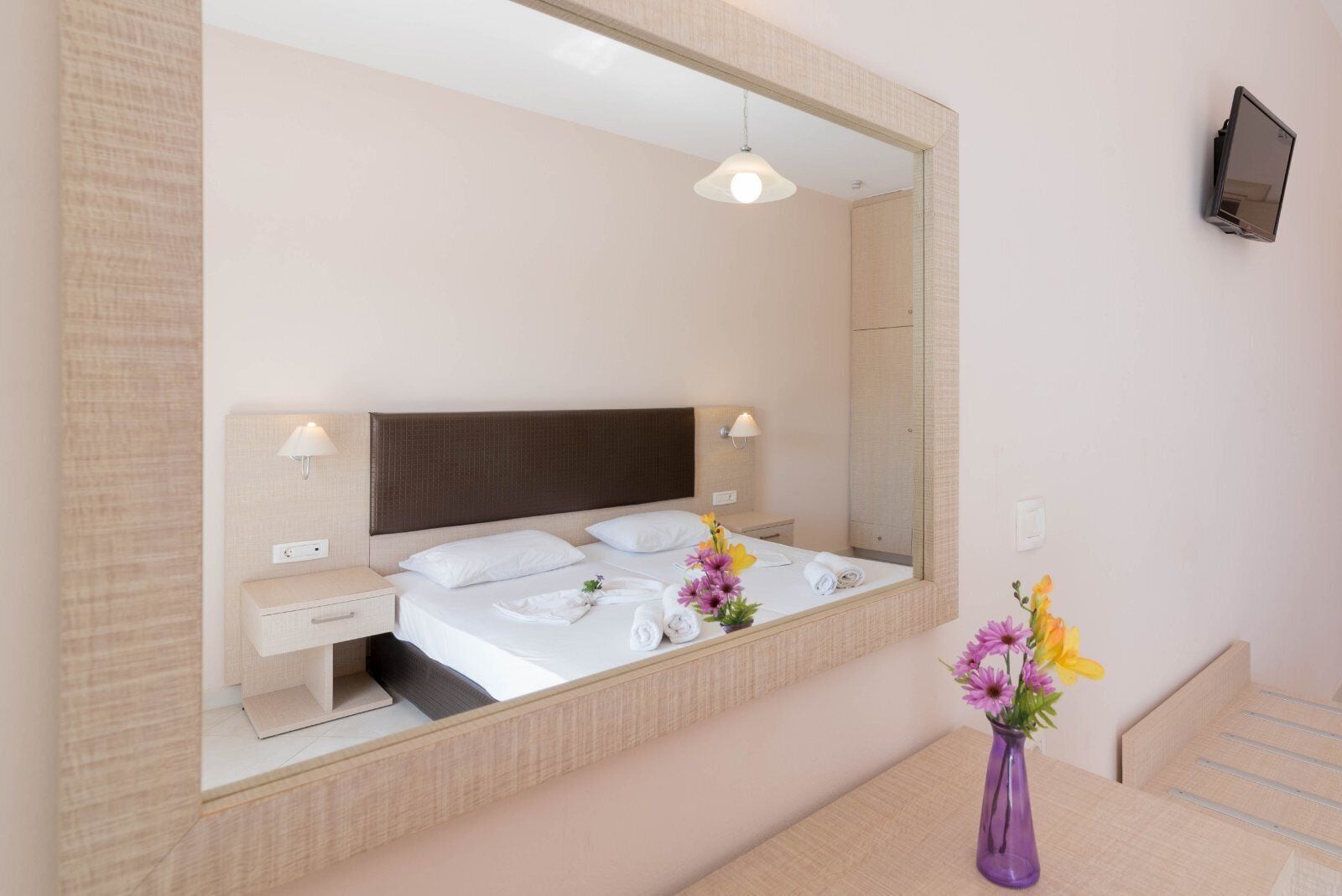 A modern hotel room with a large bed, bedside lamps, a mirror, and a small table with a vase of flowers.