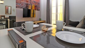 Un soggiorno moderno con un tavolo da pranzo apparecchiato per due persone, una TV a schermo piatto montata a parete e un'ampia finestra con tende.