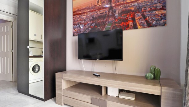 Un soggiorno moderno con una TV a schermo piatto montata a parete, un mobile basso in legno con cassetti e un dipinto raffigurante un panorama cittadino sopra la TV.