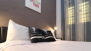 Una camera d'albergo con un letto, biancheria bianca, un cuscino nero con la scritta 'New York taxi' e un cartello 'New York' incorniciato sulla parete.