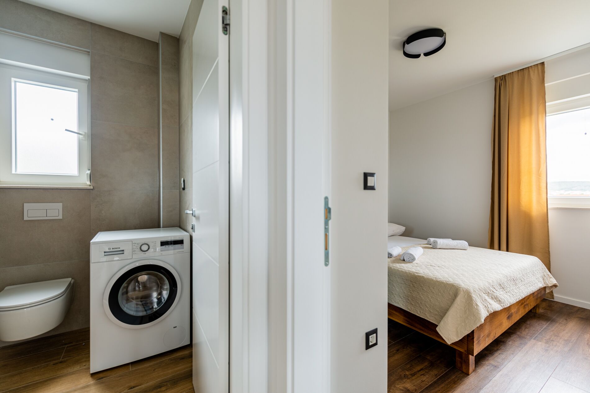 A compact room with a washing machine, a toilet, a bed, and a window with curtains.