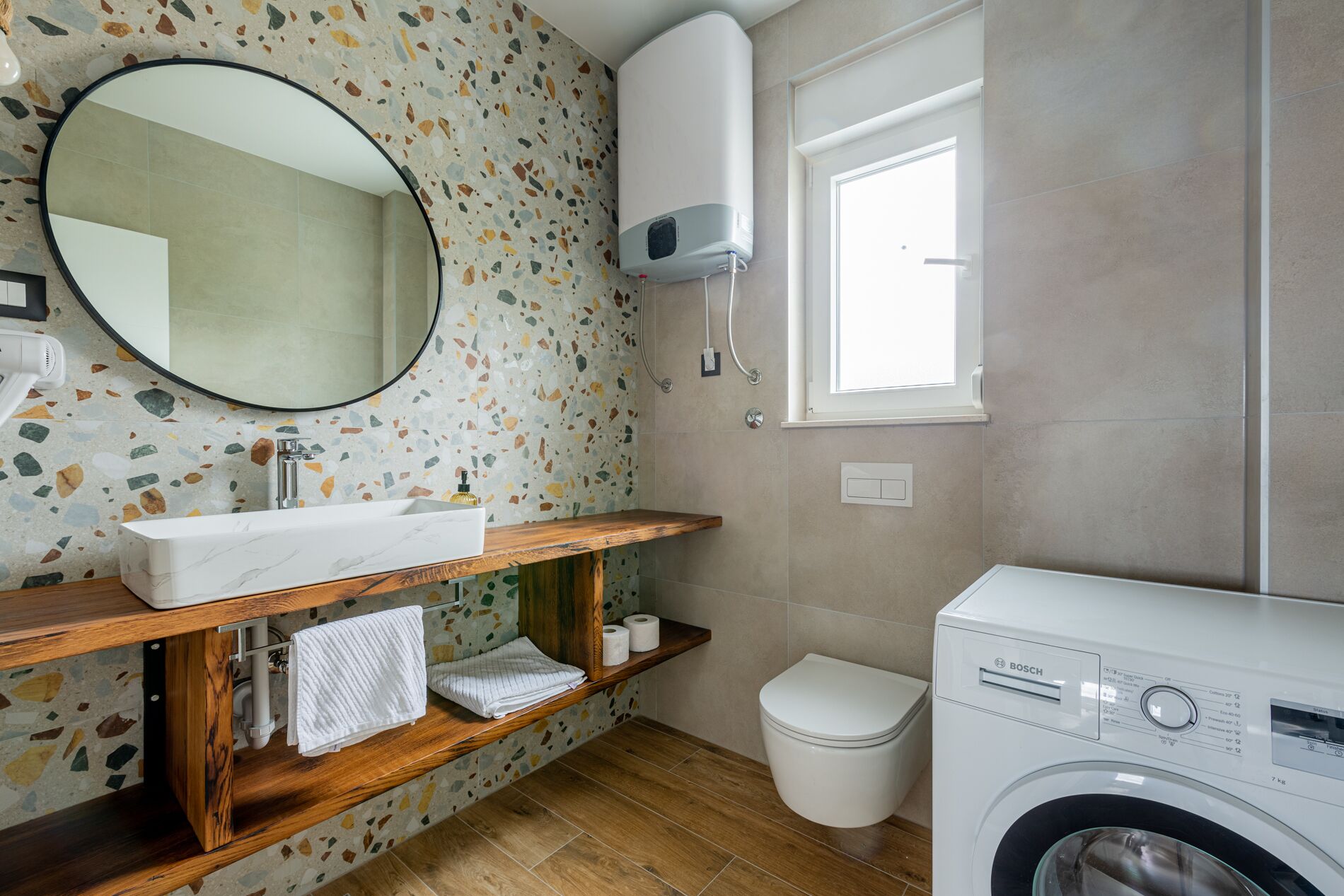 A bathroom with a white sink, wooden vanity, a round mirror, a white toilet, and a Bosch washing machine.