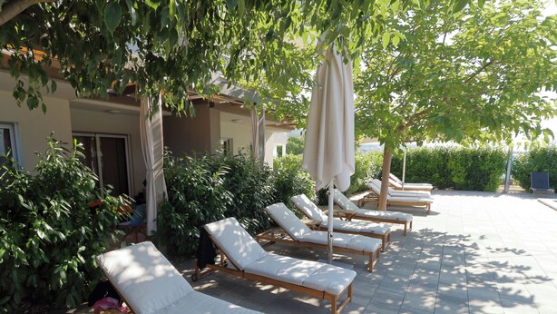 Une rangée de fauteuils de jardin en bois, dotés de coussins blancs, est disposée dans un espace extérieur ensoleillé, dont le sol est recouvert de carreaux, et est entourée de verdure. Un bâtiment se dresse en arrière-plan.