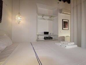 A hotel room with a neatly made bed, a hanging light fixture, a wardrobe with hanging clothes, a mirror, and a framed picture on the wall.