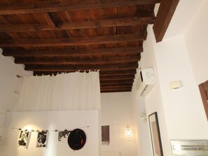 Une pièce avec un plafond en bois, des murs blancs et un rideau blanc. On y trouve des photographies en noir et blanc accrochées au mur, un chapeau noir orné d’une bordure rouge et un luminaire suspendu.