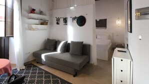 Een compacte slaapkamer met een grijze bank, een klein nachtkastje, een bed met wit beddengoed, en een wandplank met persoonlijke spullen.