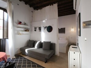 Une chambre à coucher de taille modeste, avec un canapé gris, une petite table de chevet, un lit avec une literie blanche et une étagère murale où sont exposés des objets personnels.