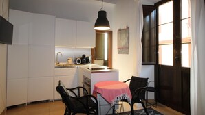 Een compacte keuken met witte kastjes, een klein eethoekje met twee stoelen en een tafel, en een groot raam dat voor natuurlijk licht zorgt.