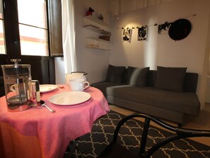 A cozy living room with a grey sofa, a coffee table with a pink tablecloth, and a dining area with a white table set for two.