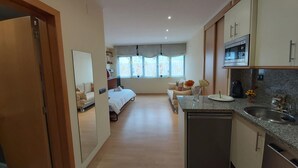 Ett kompakt hotellrum med ett litet pentry, en säng, en liten sittgrupp och ett fönster med gardiner.