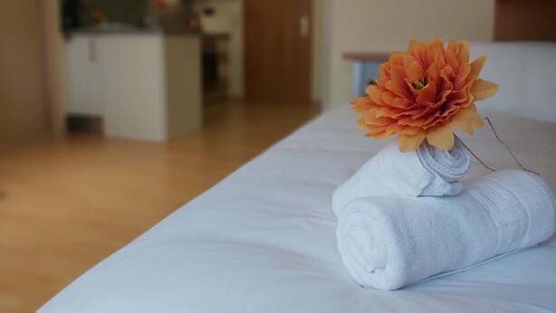 En snyggt bäddad säng med en hoprullad vit handduk och en stor orange blomma.