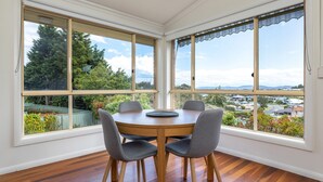 Una sala de estar con ventanales, con una mesa redonda de madera y cuatro sillas grises, que ofrece vistas a una zona residencial y árboles.